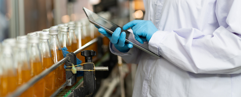 person in lab coat looking at a tablet in front of bottle filled with liquid manufacturing