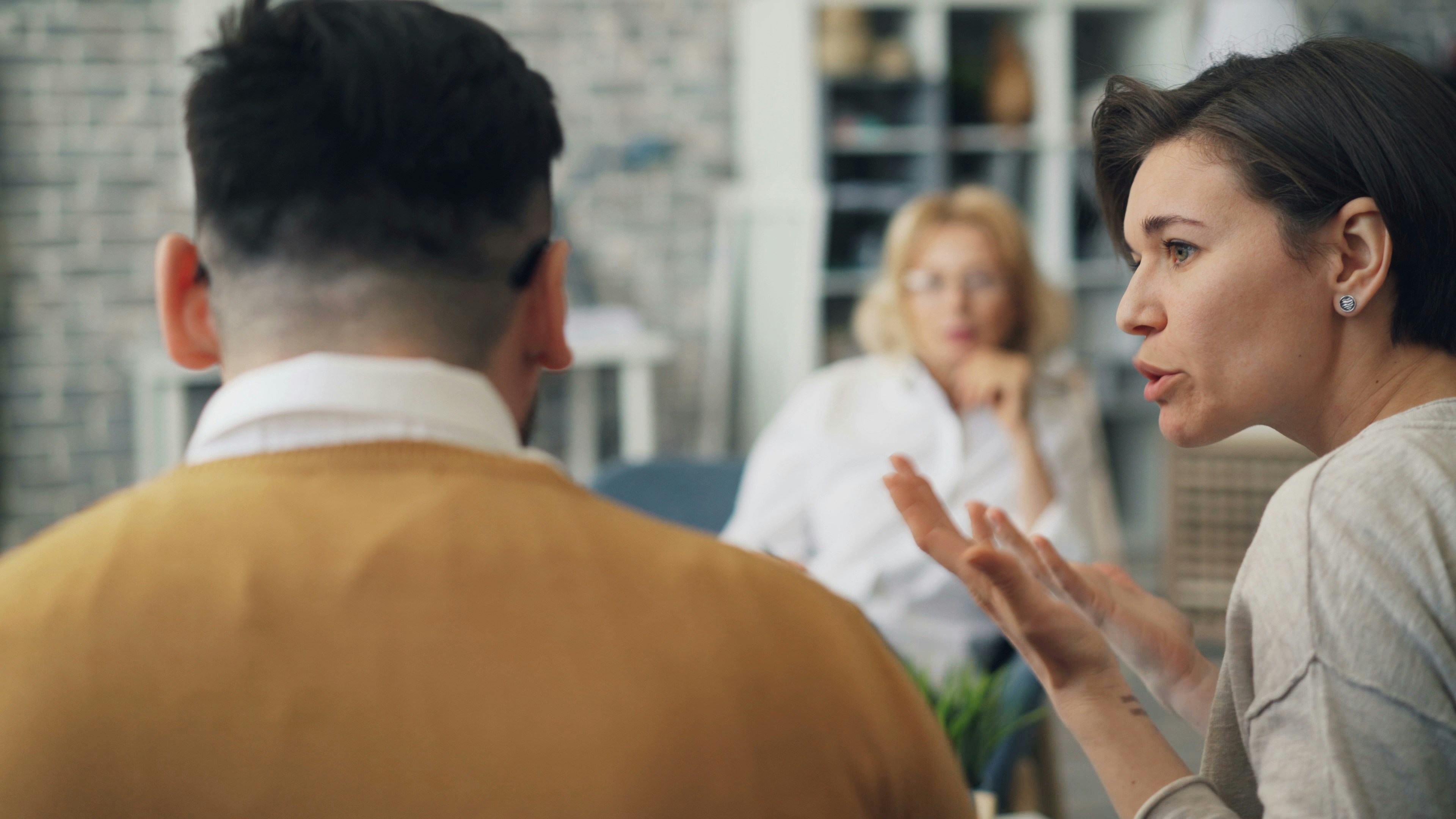 A couple in a therapy session, with the woman speaking and gesturing while the man's back is to the camera.