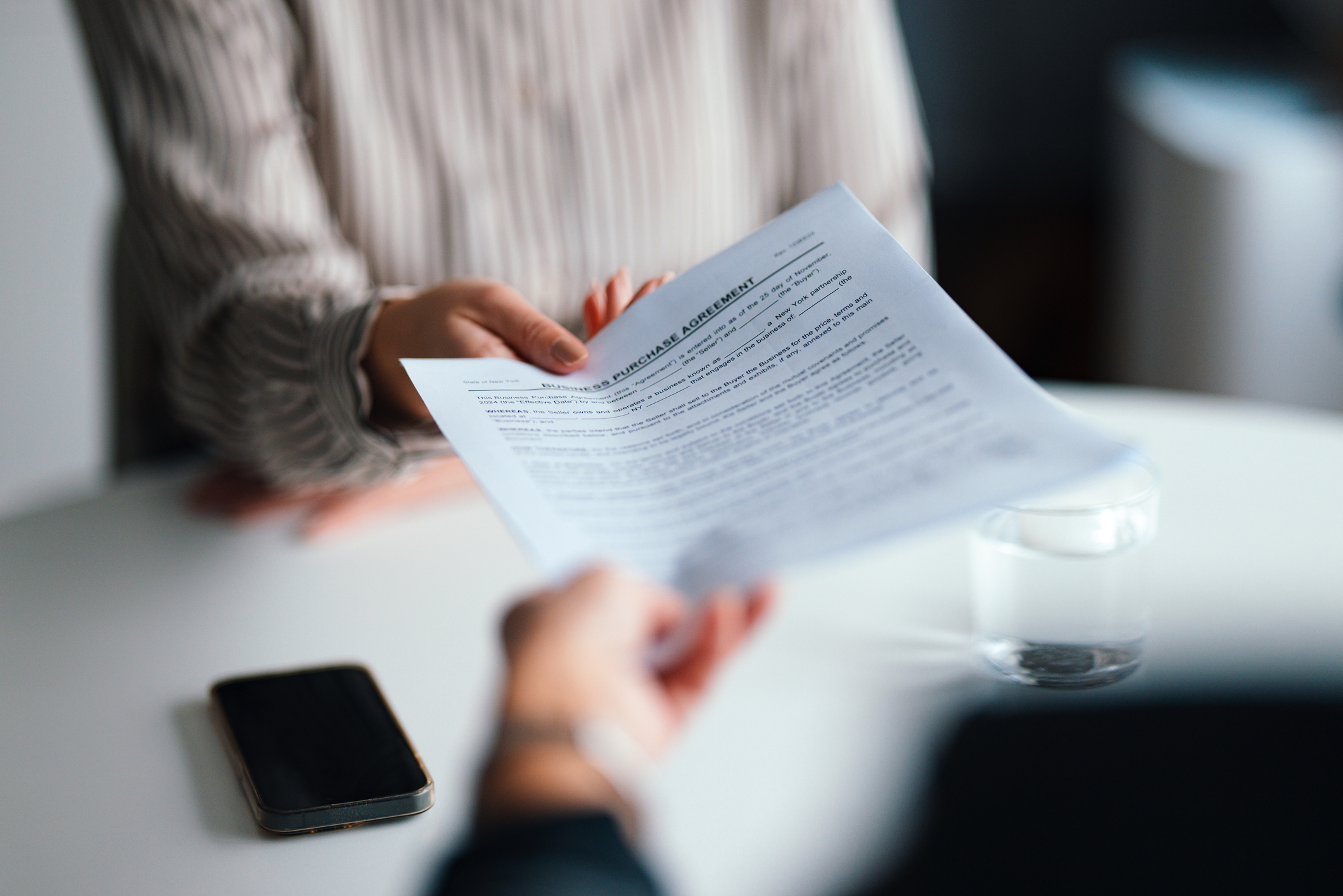 A document titled "Business Purchase Agreement" is being handed from one person to another across a table.