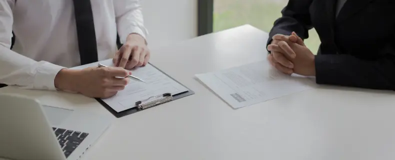 A man points to a document while interviewing a woman across a white desk.