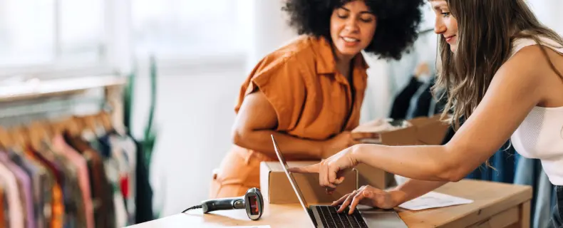 two women looking at a laptop in a store on a desk with clothing racks in the background retail