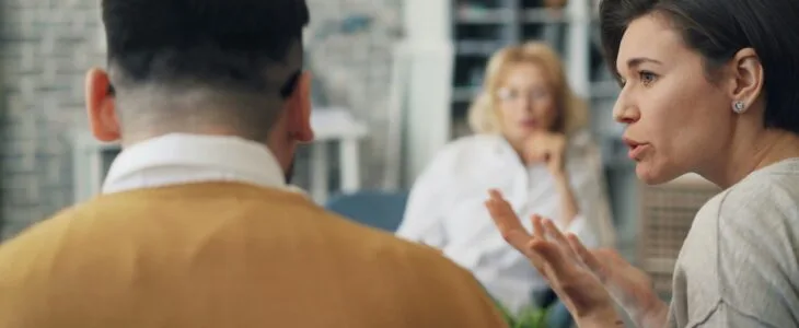 A couple in a therapy session, with the woman speaking and gesturing while the man's back is to the camera.