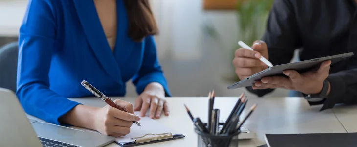 "Two professionals collaborating at an office desk, one taking notes on a clipboard and the other using a stylus on a digital tablet."